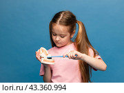 Little girl holding a model of a jaw with teeth and a toothbrush in her hands to demonstrate proper tooth brushing on blue background. Children's dentistry. Children's teeth care. Copy space. Стоковое фото, фотограф Ирина Михайличенко / Фотобанк Лори