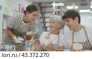 A cheerful elderly woman, along with a young girl and a guy wearing aprons, are engaged in ceramic dishware painting during a pottery class. This is a creative hobby and an intergenerational communication concept. Стоковое видео, видеограф Яков Филимонов / Фотобанк Лори