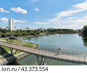 Пешеходный мост на набережной вокруг городского пруда в Зеленограде. По мосту идут две девушки. На фоне городской парк Победы, голубое небо, белые облака (2024 год). Стоковое фото, фотограф Сергей Никифоров-Гордеев / Фотобанк Лори