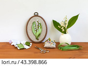 Handmade embroidery with satin ribbons of spring  lilies of the valley in hoops, bouquet of flowers in vase on wooden table. Nearby is bobbins of satin ribbons, needles and scissors for work. DIY concept. Стоковое фото, фотограф Виктория Катьянова / Фотобанк Лори