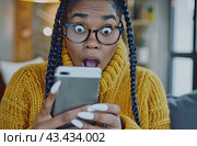 A young black woman with glasses and a surprised expression keeps a mobile phone at home. Стоковое AI-изображение, AI-художник Ярославский Максим / Фотобанк Лори
