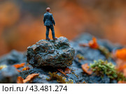 A miniature man in a business suit standing on top of a rock. The background of the image is blurred, with shades of autumn colors, which emphasizes the contrast between the person and the environment. This miniature character conveys the impression of determination and willingness to overcome obstacles, symbolizing determination and fortitude. Стоковое AI-изображение, AI-художник Анна Кривицкая / Фотобанк Лори