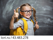 A cute girl with glasses shows the class with her hand. The child goes to school for the first time. A child with a school bag and a book. A child in the classroom on the background of a blackboard. Back to school. Стоковое AI-изображение, AI-художник Photojuli / Фотобанк Лори