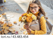 A pretty little girl with glasses and a bouquet of autumn flowers goes to school for the first time. A child with a school bag, a book and flowers. A child on a city street. Back to school. Стоковое AI-изображение, AI-художник Photojuli / Фотобанк Лори