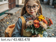 A pretty little girl with glasses and a bouquet of autumn flowers goes to school for the first time. A child with a school bag, a book and flowers. A child on a city street. Back to school. Стоковое AI-изображение, AI-художник Photojuli / Фотобанк Лори