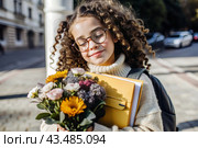 A cute little curly-haired girl with glasses and a bouquet of autumn flowers goes to school for the first time. A child with a school bag, a book and flowers. A child on a city street. Back to school. Стоковое AI-изображение, AI-художник Photojuli / Фотобанк Лори