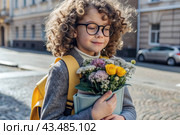 A cute little curly-haired girl with glasses and a bouquet of autumn flowers goes to school for the first time. A child with a school bag, a book and flowers. A child on a city street. Back to school. Стоковое AI-изображение, AI-художник Photojuli / Фотобанк Лори