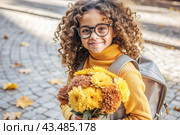 A cute little curly-haired girl with glasses and a bouquet of autumn flowers goes to school for the first time. A child with a school bag, a book and flowers. A child on a city street. Back to school. Стоковое AI-изображение, AI-художник Photojuli / Фотобанк Лори