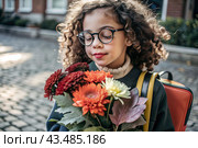 A cute little curly-haired girl with glasses and a bouquet of autumn flowers goes to school for the first time. A child with a school bag, a book and flowers. A child on a city street. Back to school. Стоковое AI-изображение, AI-художник Photojuli / Фотобанк Лори