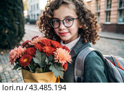 A cute little curly-haired girl with glasses and a bouquet of autumn flowers goes to school for the first time. A child with a school bag, a book and flowers. A child on a city street. Back to school. Стоковое AI-изображение, AI-художник Photojuli / Фотобанк Лори