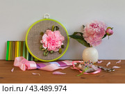Handmade embroidery with satin ribbons of peony flower in hoop, bouquet of fresh peonies in vase on wooden table. Nearby is set of colorful satin ribbons, needles and scissors for work. Front view. DIY concept. Стоковое фото, фотограф Виктория Катьянова / Фотобанк Лори