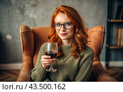 Close-up. A modern, stylish, beautiful woman with red hair and a glass of red wine in an armchair in the living room of the house. Стоковое AI-изображение, AI-художник Photojuli / Фотобанк Лори