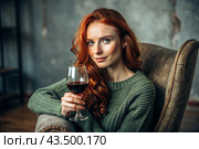 Close-up. A modern, stylish, beautiful woman with red hair and a glass of red wine in an armchair in the living room of the house. Стоковое AI-изображение, AI-художник Photojuli / Фотобанк Лори