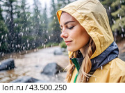 Close-up. A happy young woman in a raincoat enjoys the rain on the background of a green forest, the concept of enjoying a natural leisurely life, environment, lifestyle. Стоковое AI-изображение, AI-художник Photojuli / Фотобанк Лори