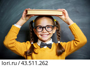A pretty girl with glasses and books on her head. The child goes to school for the first time. A child in the classroom on the background of a blackboard. Back to school. Стоковое AI-изображение, AI-художник Photojuli / Фотобанк Лори