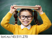 A pretty girl with glasses and books on her head. The child goes to school for the first time. A child in the classroom on the background of a blackboard. Back to school. Стоковое AI-изображение, AI-художник Photojuli / Фотобанк Лори