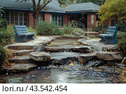 A cozy garden corner with two blue benches located on either side of a small pond with cascading waterfalls. The water flows smoothly over the stones, creating a feeling of calm and comfort. In the background you can see a brick building with large windows, surrounded by greenery and shrubs. The atmosphere of the garden is peaceful, ideal for relaxing and enjoying nature. Стоковое AI-изображение, AI-художник Анна Кривицкая / Фотобанк Лори