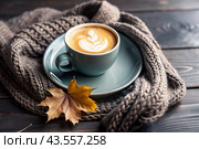 A seasonal drink. Delicious hot cappuccino in a mug with a saucer and a knitted snood, a scarf on a wooden table in the interior of the cafe. It's a rainy autumn day. Стоковое AI-изображение, AI-художник Photojuli / Фотобанк Лори