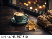 A seasonal drink. Delicious hot cappuccino in a mug with a saucer and a knitted snood, a scarf on a wooden table in the interior of the cafe. It's a rainy autumn day. Стоковое AI-изображение, AI-художник Photojuli / Фотобанк Лори