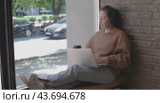 Woman in headphones sitting on cafe windowsill with laptop drinking coffee. Woman drinking coffee and embraces cafe ambiance effortlessly navigating workload in cozy setting with hot coffee in cafe. Стоковое видео, видеограф Olesia Kondrateva / Фотобанк Лори