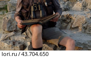 A man dressed as a researcher and with binoculars sits on the rocks and studies an old book. Стоковое видео, видеограф Константин Скуридин / Фотобанк Лори
