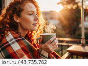 A red-haired, beautiful woman wrapped in a plaid with a mug of hot coffee on a beautiful patio in the rays of the setting sun. The girl inhales the aroma of the drink. Comfort and harmony. Стоковое AI-изображение, AI-художник Photojuli / Фотобанк Лори