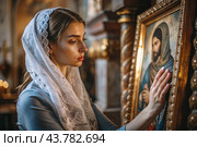 Portrait of a beautiful young woman in a headscarf in an Orthodox church. A girl prays in a church against the background of icons. Стоковое AI-изображение, AI-художник Photojuli / Фотобанк Лори