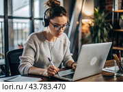 A young female freelance businessman in glasses and headphones is working on a laptop or shopping in an online store. The woman is writing something in a notebook. Стоковое AI-изображение, AI-художник Photojuli / Фотобанк Лори