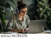 A young female freelance businessman in glasses and headphones is working on a laptop or shopping in an online store. The woman is writing something in a notebook. Стоковое AI-изображение, AI-художник Photojuli / Фотобанк Лори