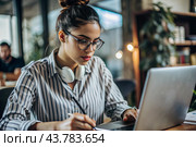 A young female freelance businessman in glasses and headphones is working on a laptop or shopping in an online store. The woman is writing something in a notebook. Стоковое AI-изображение, AI-художник Photojuli / Фотобанк Лори