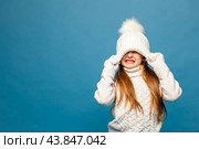 Child girl in a white sweatshirt and knitted hat with pompom on a blue background. The concept of children's emotions, joy and delight. Advertising clothing and sales. Ski resort vacation, copy space. Стоковое фото, фотограф Ирина Михайличенко / Фотобанк Лори