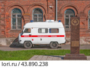 Белая машина скорой помощи стоит перед историческим зданием богадельни Мещериных (ныне центральная городская больница). Рядом памятный знак на братской могиле в память о погибших в 1919 году продотрядовцах.  Город Юрьев-Польский, улица 1 Мая, дом 16. Владимирская область, Россия (2020 год). Редакционное фото, фотограф Наталья Николаева / Фотобанк Лори