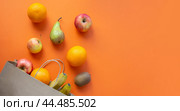 Flat lay of fruits, including an apple, pear, bananas, oranges and a kiwi, arranged around a brown paper bag on an orange background. Стоковое фото, фотограф Ольга Петрова-Апостолова / Фотобанк Лори