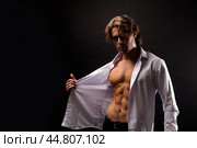 Perfect young man fashion model showing unbuttoned white shirt with abs muscle press poses in shadow at black. Portrait of stylish guy with perfect muscular sexy torso, studio shot. Copy ad text space. Стоковое фото, фотограф Aleksandr Golubev / Фотобанк Лори