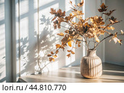 Branches with golden autumn leaves in a ceramic vase on a wooden table by the window. The concept of home comfort. Autumn decor for Halloween. Стоковое AI-изображение, AI-художник Photojuli / Фотобанк Лори