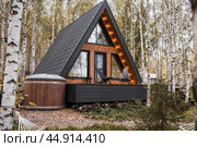 A-shaped frame house in Scandinavian style. The landscape. Quiet rest. The concept of glamping and chalet rentals. A wooden house with a veranda in nature around an autumn birch forest. Стоковое фото, фотограф Photojuli / Фотобанк Лори