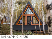 A-shaped frame house in Scandinavian style. The landscape. Quiet rest. The concept of glamping and chalet rentals. A wooden house with a veranda in nature around an autumn birch forest. Стоковое фото, фотограф Photojuli / Фотобанк Лори