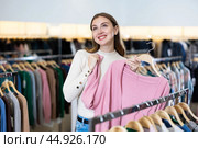Focused young woman holding a pink cardigan on a rack while choosing clothes in a showroom. Стоковое фото, фотограф Яков Филимонов / Фотобанк Лори