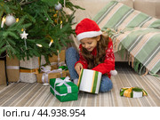 Christmas spirit. Happy little smiling girl opening gift box sitting on the floor near the Christmas tree. Happy girl unwrapping christmas present looking in box. Holidays and celebrations concept. Стоковое фото, фотограф Ирина Михайличенко / Фотобанк Лори