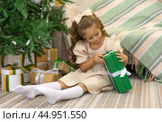 Christmas spirit. Happy little smiling girl opening gift box sitting on the floor near the Christmas tree. Happy girl unwrapping Christmas present looking in box. Holidays and celebrations concept. Стоковое фото, фотограф Ирина Михайличенко / Фотобанк Лори