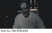 A pale-faced creature with sunglasses and a wide grinning mouth. An attic monster in a white sweatshirt and baseball cap in a dim room with rays of light coming through the cracks. Стоковое видео, видеограф Воловодов Владимир / Фотобанк Лори