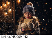 A little girl in winter clothes holding an envelope outside in snowy weather against the background of lights. A letter for Santa Claus. The winter atmosphere of a festive invitation. Стоковое AI-изображение, AI-художник Photojuli / Фотобанк Лори