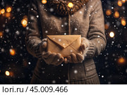 A little girl in winter clothes holding an envelope outside in snowy weather against the background of lights. A letter for Santa Claus. The winter atmosphere of a festive invitation. Стоковое AI-изображение, AI-художник Photojuli / Фотобанк Лори