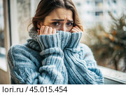 Winter cold. A young woman in a warm knitted blue sweater with a high neck at home. He looks at the camera, covering his sad face. Depression, sadness, loneliness, social phobia. Стоковое AI-изображение, AI-художник Photojuli / Фотобанк Лори
