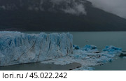 A fragment of a melting glacier. A wall of cracked blue ice rises above a turquoise glacial lake. Melted ice floes float in the water. Mountains in fog and clouds. Perito Moreno. El Calafate.Argentina. Стоковое фото, фотограф Вера Ивановна Кубарева / Фотобанк Лори