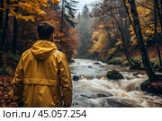 A man in a yellow rubber raincoat on the background of a huge river in a forest in the mountains. The guy travels and enjoys life and the environment. Tourism. Стоковое AI-изображение, AI-художник Photojuli / Фотобанк Лори