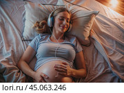 A pregnant woman with headphones on, lying in bed. The influence of music on pregnancy. A woman is preparing to become a mother. Pregnancy and childbirth. Стоковое AI-изображение, AI-художник Photojuli / Фотобанк Лори