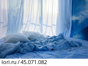 Unmade bed with blue sheets near window covered by sheer white curtains. Стоковое AI-изображение, AI-художник Иван Рябоконь / Фотобанк Лори