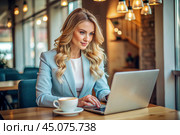 A blonde businesswoman in a cafe. She is wearing a casual blue jacket. A woman is working on a laptop, next to a cup of coffee. The businesswoman looks at the computer screen. Стоковое AI-изображение, AI-художник Photojuli / Фотобанк Лори