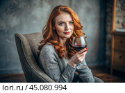 Close-up. A modern, stylish, beautiful woman with red hair and a glass of red wine in an armchair in the living room of the house. Стоковое AI-изображение, AI-художник Photojuli / Фотобанк Лори